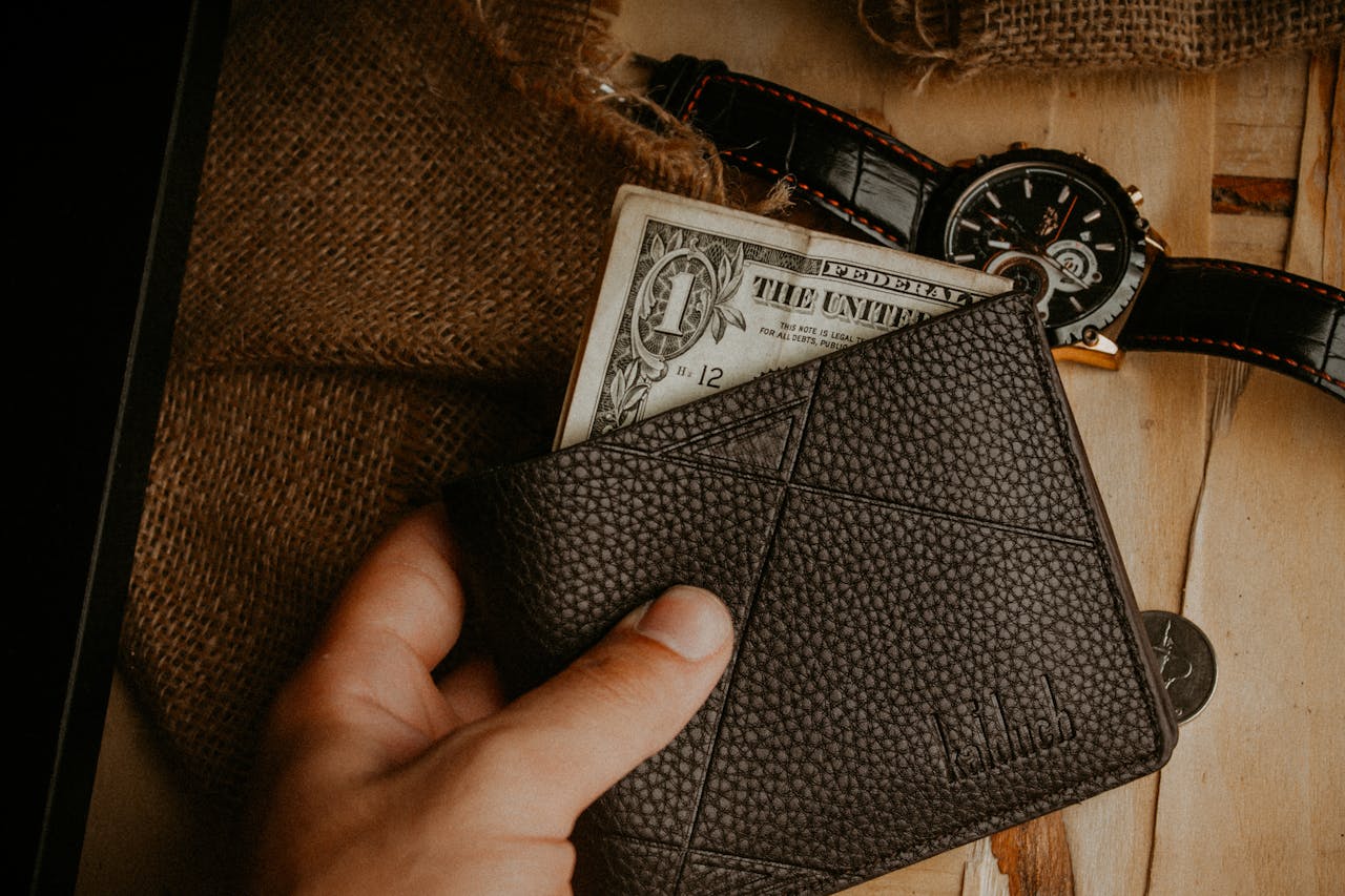 A hand holds a leather wallet with a dollar bill exposed, next to a wristwatch on a wooden surface.
